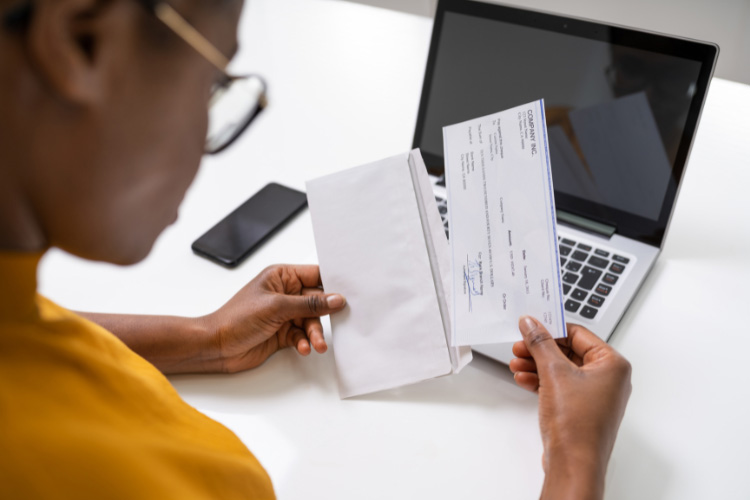 A woman sits in front of a laptop and phone, holding and examining an envelope and paycheck.