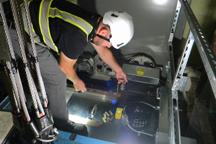 A man in a hard hat and high-visibility vest stands on top of an elevator cab, closing the hatch. 