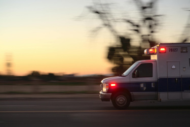 An ambulance with flashing lights driving on a road, against a blurred background.