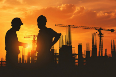 Silhouettes of two construction workers standing in front of a construction site against an orange sunset